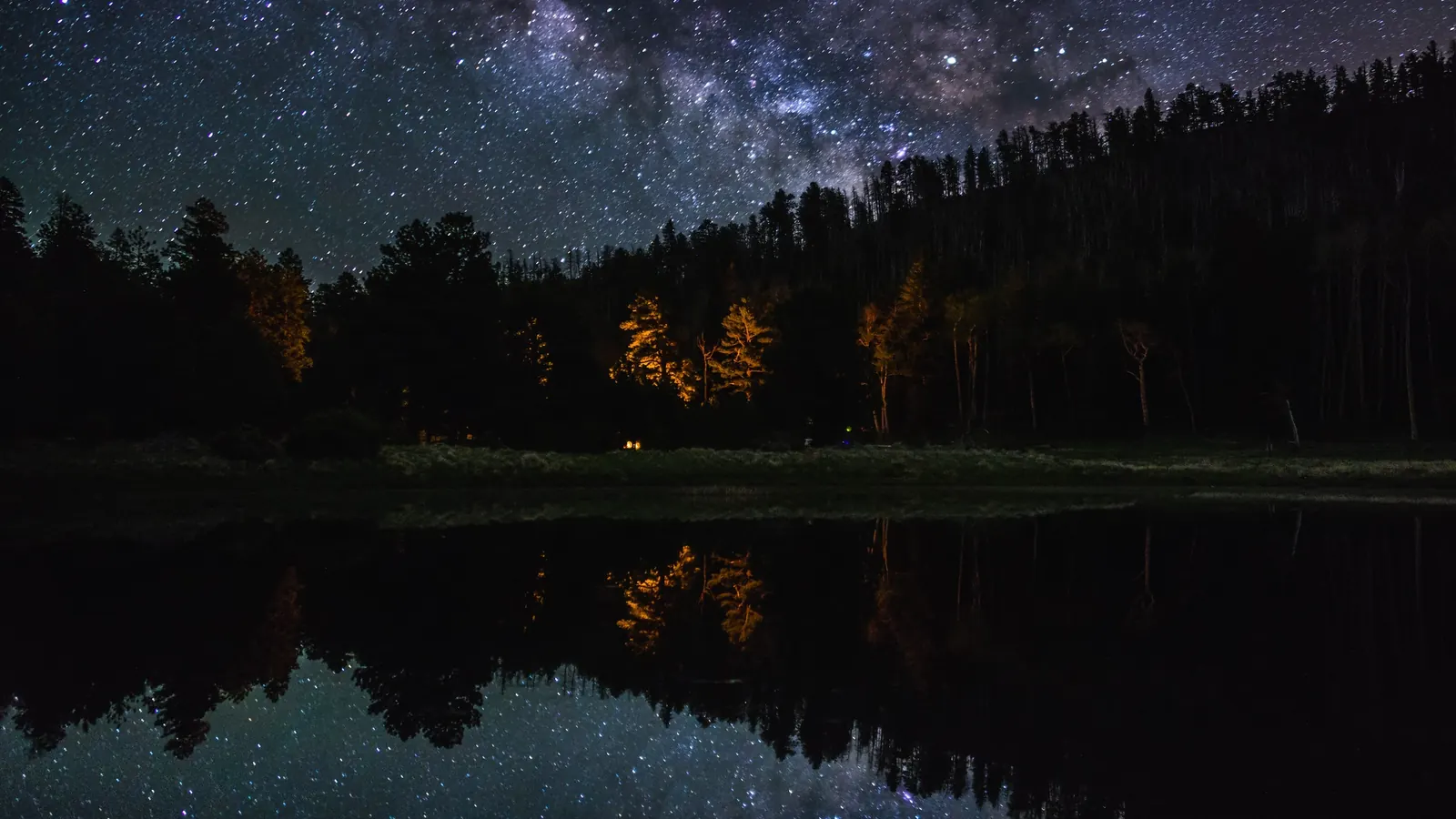The Milky Way over a forest and a still mountain lake at night
