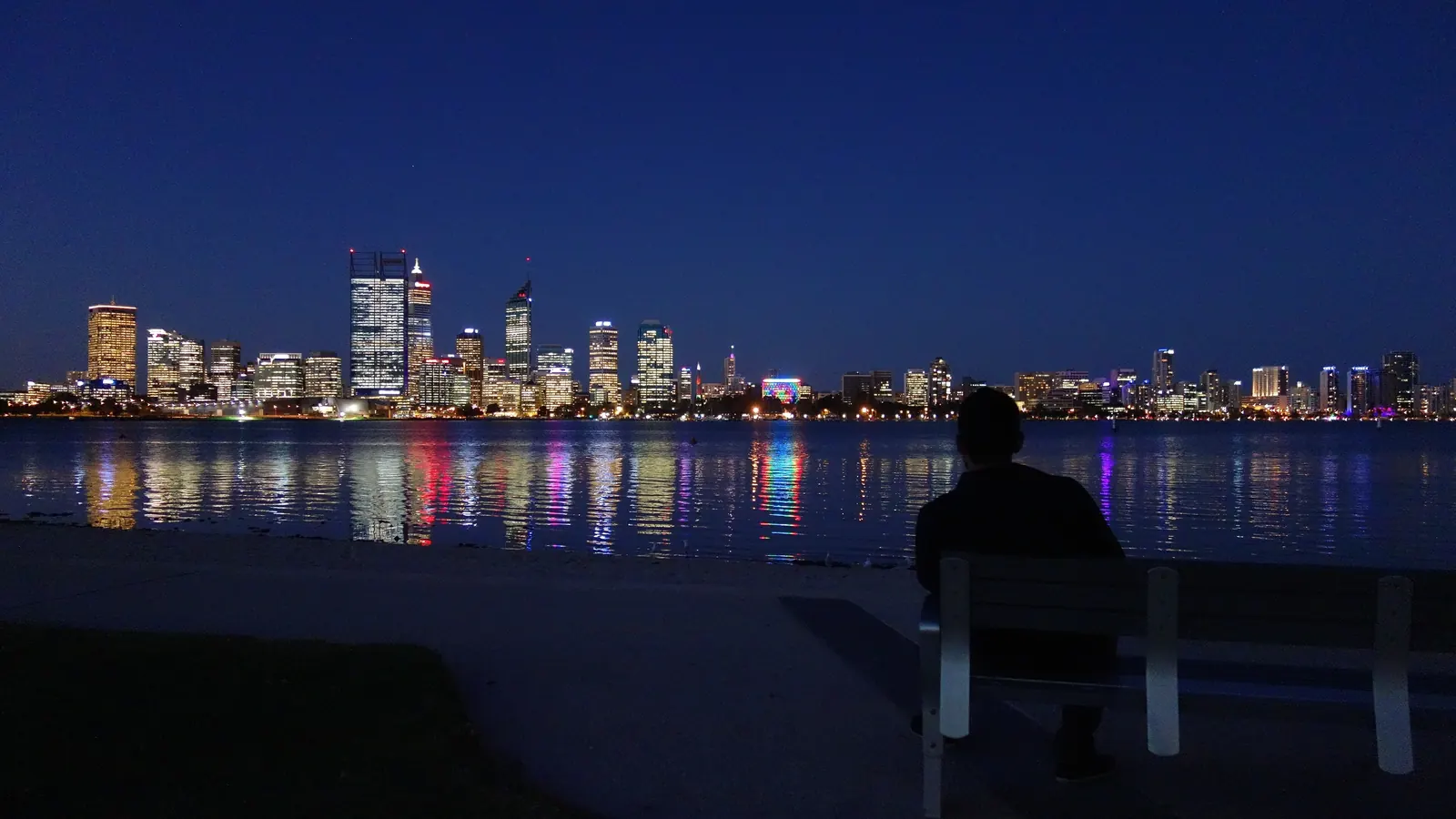 A person on a bench looking across the water at a city skyline lit up at night