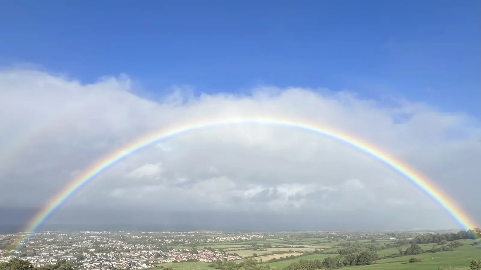 A full rainbow arching over green countryside and a distant town